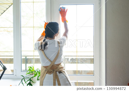 Woman in rubber gloves and apron manually washes window balcony of house with rag cleaner and mop inside interior. Restoring order and cleanliness in spring, cleaning servise Woman in rubber gloves and apron manually washes window balcony of house with rag cleaner and mop inside interior. Restoring order and cleanliness in spring, cleaning servise 130994872