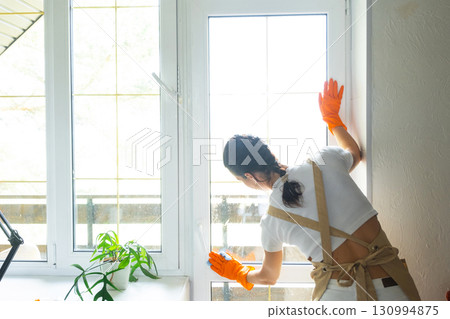 Woman in rubber gloves and apron manually washes window balcony of house with rag cleaner and mop inside interior. Restoring order and cleanliness in spring, cleaning servise Woman in rubber gloves and apron manually washes window balcony of house with rag cleaner and mop inside interior. Restoring order and cleanliness in spring, cleaning servise 130994875