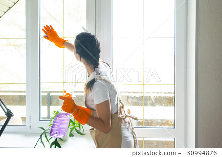 Woman in rubber gloves and apron manually washes window balcony of house with rag cleaner and mop inside interior. Restoring order and cleanliness in spring, cleaning servise 130994876