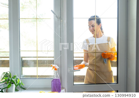 Woman in rubber gloves and apron manually washes window balcony of house with rag cleaner and mop outside behind glass. Restoring order and cleanliness in spring, cleaning servise Woman in rubber gloves and apron manually washes window balcony of house with rag cleaner and mop outside behind glass. Restoring order and cleanliness in spring, cleaning servise 130994910