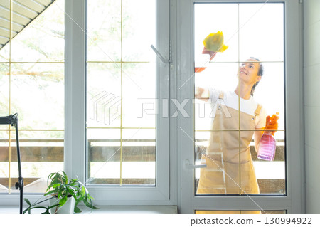 Woman in rubber gloves and apron manually washes window balcony of house with rag cleaner and mop outside behind glass. Restoring order and cleanliness in spring, cleaning servise 130994922