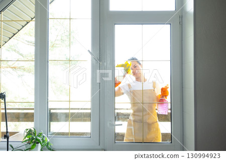 Woman in rubber gloves and apron manually washes window balcony of house with rag cleaner and mop outside behind glass. Restoring order and cleanliness in spring, cleaning servise Woman in rubber gloves and apron manually washes window balcony of house with rag cleaner and mop outside behind glass. Restoring order and cleanliness in spring, cleaning servise 130994923
