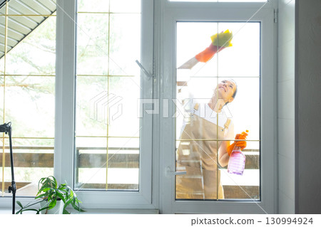 Woman in rubber gloves and apron manually washes window balcony of house with rag cleaner and mop outside behind glass. Restoring order and cleanliness in spring, cleaning servise Woman in rubber gloves and apron manually washes window balcony of house with rag cleaner and mop outside behind glass. Restoring order and cleanliness in spring, cleaning servise 130994924