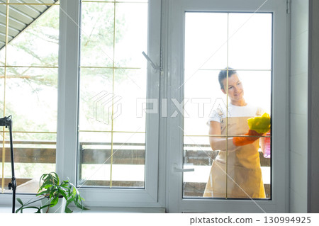 Woman in rubber gloves and apron manually washes window balcony of house with rag cleaner and mop outside behind glass. Restoring order and cleanliness in spring, cleaning servise Woman in rubber gloves and apron manually washes window balcony of house with rag cleaner and mop outside behind glass. Restoring order and cleanliness in spring, cleaning servise 130994925