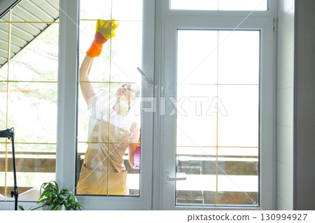 Woman in rubber gloves and apron manually washes window balcony of house with rag cleaner and mop outside behind glass. Restoring order and cleanliness in spring, cleaning servise Woman in rubber gloves and apron manually washes window balcony of house with rag cleaner and mop outside behind glass. Restoring order and cleanliness in spring, cleaning servise 130994927