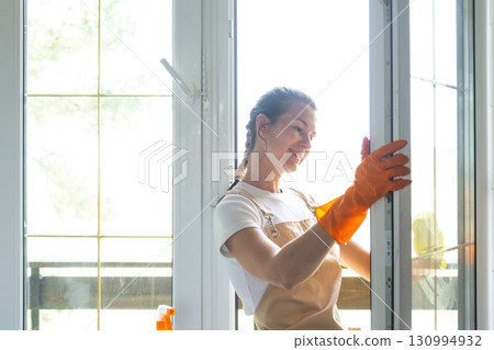 Woman in rubber gloves and apron manually washes window balcony of house with rag cleaner and mop inside interior. Restoring order and cleanliness in spring, cleaning servise 130994932