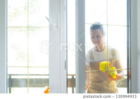 Woman in rubber gloves and apron manually washes window balcony of house with rag cleaner and mop outside behind glass. Restoring order and cleanliness in spring, cleaning servise 130994933