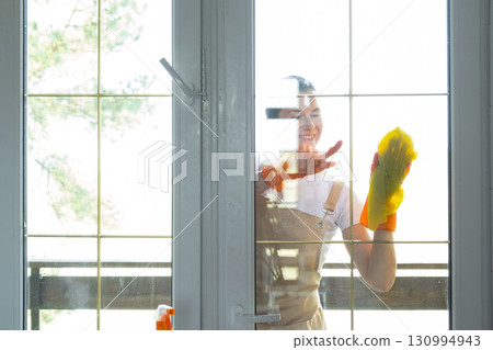 Woman in rubber gloves and apron manually washes window balcony of house with rag cleaner and mop outside behind glass. Restoring order and cleanliness in spring, cleaning servise 130994943