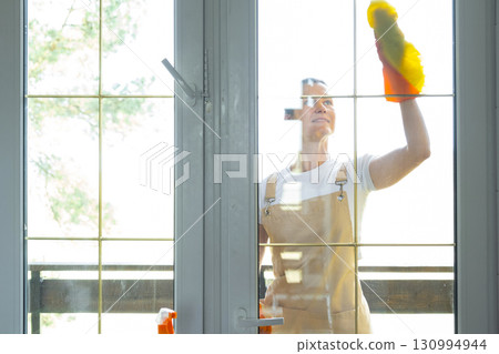 Woman in rubber gloves and apron manually washes window balcony of house with rag cleaner and mop outside behind glass. Restoring order and cleanliness in spring, cleaning servise Woman in rubber gloves and apron manually washes window balcony of house with rag cleaner and mop outside behind glass. Restoring order and cleanliness in spring, cleaning servise 130994944