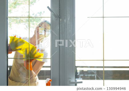 Woman in rubber gloves and apron manually washes window balcony of house with rag cleaner and mop outside behind glass. Restoring order and cleanliness in spring, cleaning servise 130994946