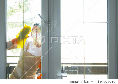 Woman in rubber gloves and apron manually washes window balcony of house with rag cleaner and mop outside behind glass. Restoring order and cleanliness in spring, cleaning servise Woman in rubber gloves and apron manually washes window balcony of house with rag cleaner and mop outside behind glass. Restoring order and cleanliness in spring, cleaning servise 130994949