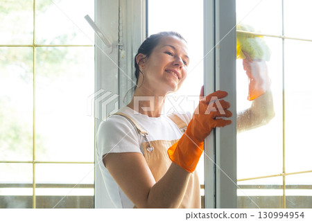 Woman in rubber gloves and apron manually washes window balcony of house with rag cleaner and mop outside behind glass. Restoring order and cleanliness in spring, cleaning servise 130994954