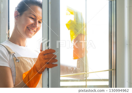 Woman in rubber gloves and apron manually washes window balcony of house with rag cleaner and mop outside behind glass. Restoring order and cleanliness in spring, cleaning servise Woman in rubber gloves and apron manually washes window balcony of house with rag cleaner and mop outside behind glass. Restoring order and cleanliness in spring, cleaning servise 130994962