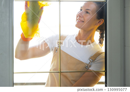Woman in rubber gloves and apron manually washes window balcony of house with rag cleaner and mop outside behind glass. Restoring order and cleanliness in spring, cleaning servise 130995077