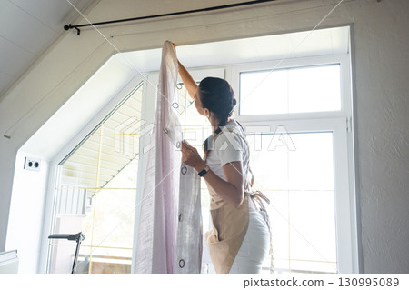 Woman in apron hangs transparent tulle curtains on large attic windows in the house inside the white interior. Spring cleaning, tidying up Woman in apron hangs transparent tulle curtains on large attic windows in the house inside the white interior. Spring cleaning, tidying up 130995089