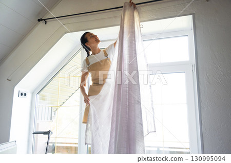 Woman in apron hangs transparent tulle curtains on large attic windows in the house inside the white interior. Spring cleaning, tidying up 130995094