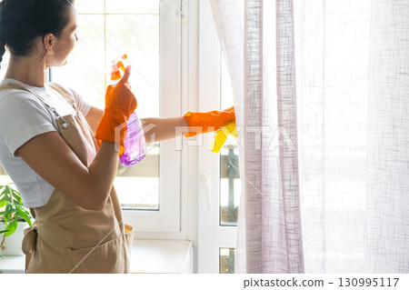 Woman in rubber gloves and apron manually washes window balcony of house with rag cleaner and mop inside interior. Restoring order and cleanliness in spring, cleaning servise Woman in rubber gloves and apron manually washes window balcony of house with rag cleaner and mop inside interior. Restoring order and cleanliness in spring, cleaning servise 130995117