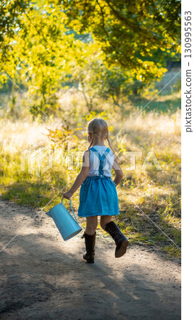 Little girl in blue denim dress and boots walking with watering can on forest path 130995563