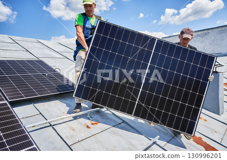 Workers building solar panel system on metal rooftop of house. Two men installers carrying photovoltaic solar module outdoors. Alternative, green and renewable energy generation concept. 130996201