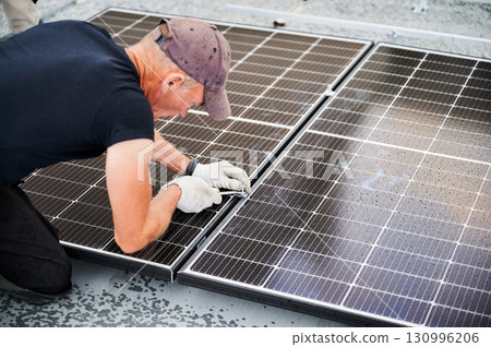Worker building photovoltaic solar panel system on rooftop of house. Close up of man engineer in gloves installing solar module with help of wrench outdoors. Alternative and renewable energy concept. 130996206