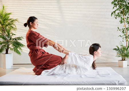 Masseuse making thai yoga massage. Woman therapist doing traditional massage treatment, stretching female patient's body. Client lying on mat, enjoying hand massage in calm atmosphere. 130996207
