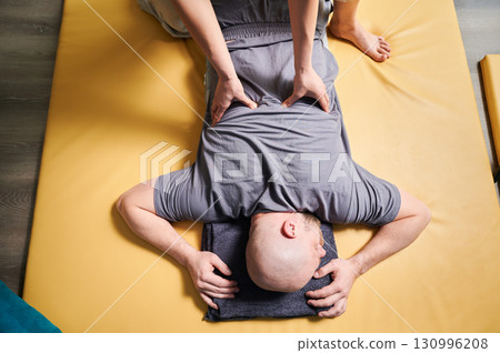 Masseuse making thai yoga massage. Therapist doing traditional massage treatment for male patient indoors. Bald client lying on mat, meditating, enjoying hand massage in calm atmosphere. 130996208