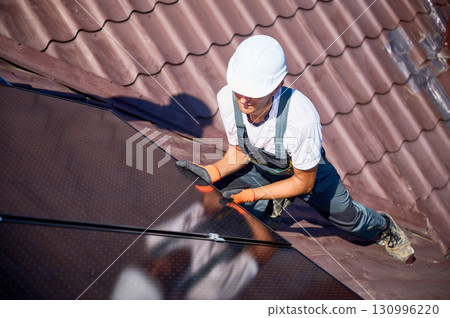 Worker building solar panel system on rooftop of house. Man engineer in helmet installing photovoltaic solar module outdoors. Alternative, green and renewable energy generation concept. 130996220