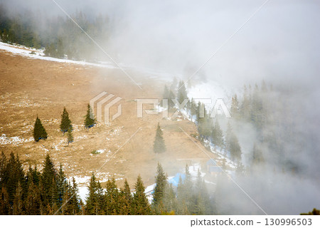 Small cabin nestled in clearing on mountainside, surrounded by dense pine forests. Scene shrouded in mist, creating a tranquil and mysterious atmosphere. Snow patches linger on the ground. Small cabin nestled in clearing on mountainside, surrounded by dense pine forests. Scene shrouded in mist, creating a tranquil and mysterious atmosphere. Snow patches linger on the ground. 130996503