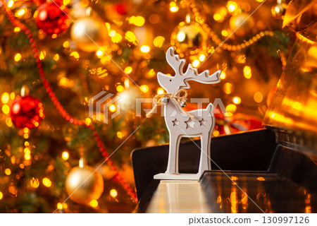 Deer toy on piano with Christmas tree with bright lights on the background. Christmas atmosphere Deer toy on piano with Christmas tree with bright lights on the background. Christmas atmosphere 130997126