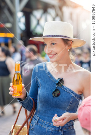 Smiling young adult happy woman wear denim outfit straw hat hold little bright orange alcohol beverage bottle standing outdoors. Sunglasses hang from her neckline enjoys casual summer city atmosphere 130997649