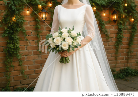 Classic bride holding white rose bouquet next to brick wall decorated with lanterns and greenery. concept of wedding elegance, bridal beauty, timeless romance. 130999475