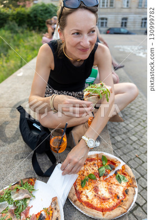 Tourist woman enjoys pizza and Aperol Spritz while sitting outdoors in Berlin during summer day. Relaxed street food atmosphere highlights casual travel lifestyle with authentic culinary experience Tourist woman enjoys pizza and Aperol Spritz while sitting outdoors in Berlin during summer day. Relaxed street food atmosphere highlights casual travel lifestyle with authentic culinary experience 130999782