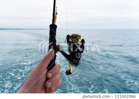 Hands of a fisherman with a spinning rod with the line with a line on a motor boat in the sea. Fishing rod and hands of fisherman over ocean water surface. Hands of a fisherman with a spinning rod with the line with a line on a motor boat in the sea. Fishing rod and hands of fisherman over ocean water surface. 131002066