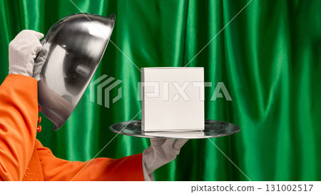 Waiter hand lifting silver cloche to reveal white box on tray against green curtain backdrop 131002517