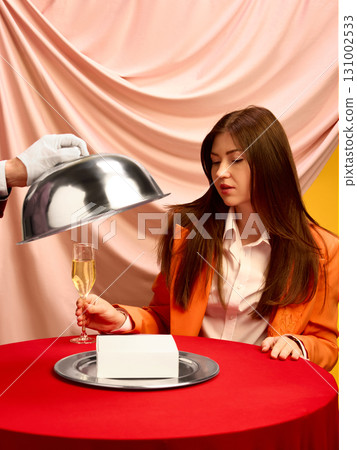 Woman with champagne watching cloche reveal white copy space box on red table with pink backdrop 131002533