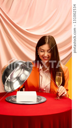 Smiling woman with champagne and cloche unveiling mockup box on red table with pink fabric 131002534