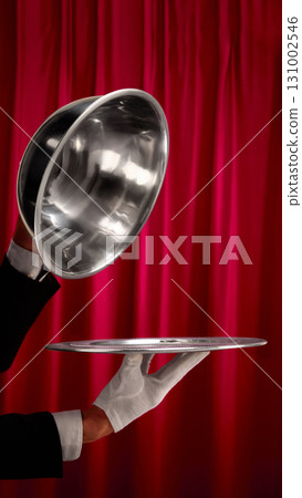 Silver cloche lifted by waiter hand with white glove against red curtain background 131002546