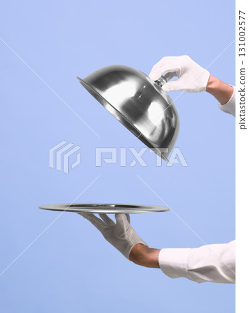 Hands lifting silver cloche from tray on blue background. Negative space for ads Hands lifting silver cloche from tray on blue background. Negative space for ads 131002577