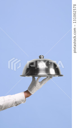 Waiter hand with silver cloche on blue background Waiter hand with silver cloche on blue background 131002578