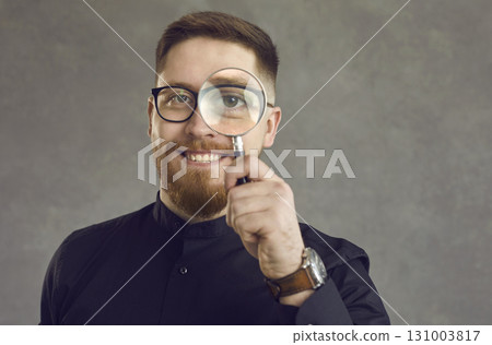 Positive young detective man looking through a magnifying glass while standing on gray background. Positive young detective man looking through a magnifying glass while standing on gray background. 131003817
