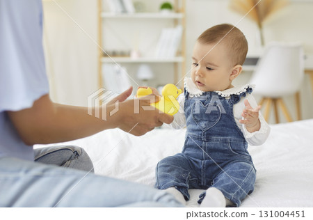 Baby sitting on bed reaching for yellow rubber duck offered by mom in soft light Baby sitting on bed reaching for yellow rubber duck offered by mom in soft light 131004451