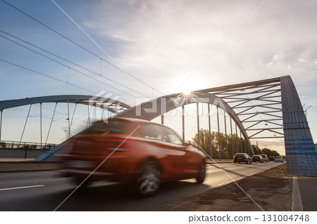 Scenic dynamic view of Magdeburg steel arch Jerusalem bridge cars crossing under warm sunrise sunlight. Urban composition highlights architectural structure traffic movement modern city atmosphere 131004748
