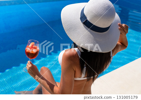 Beautiful brunette woman in a hat sitting and enjoying cocktail at swimming pool 131005179