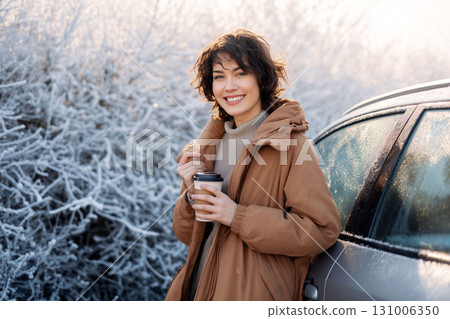 Smiling woman in warm coat holding coffee cup outdoors in frosty winter morning near frozen car Smiling woman in warm coat holding coffee cup outdoors in frosty winter morning near frozen car 131006350