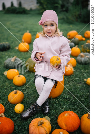 A young girl sits among pumpkins in a grassy field during autumn, happily inspecting a pumpkin A young girl sits among pumpkins in a grassy field during autumn, happily inspecting a pumpkin 131006524