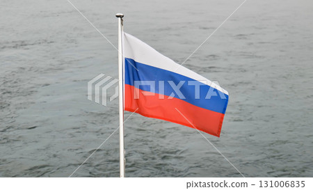 Russian flag waving vigorously, billowing against muted grey waterscape, symbolizing national spirit and patriotic sentiment 131006835