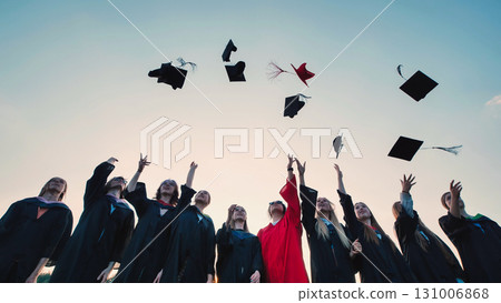 Group of cheerful graduates celebrating their academic achievement by tossing their graduation caps into the air against a sunset sky 131006868