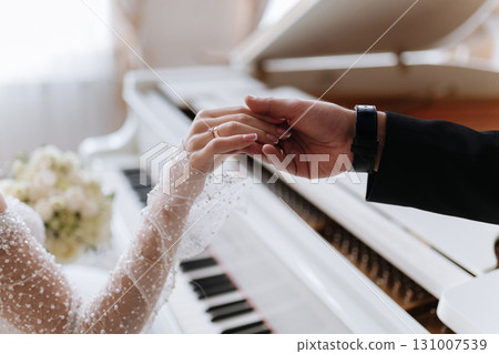 Elegant wedding moment of bride and groom gently holding hands near white piano Elegant wedding moment of bride and groom gently holding hands near white piano 131007539