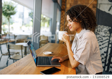 A Young Woman is Enjoying Her Coffee While Working on Her Laptop in a Cozy Cafe Setting 131008529