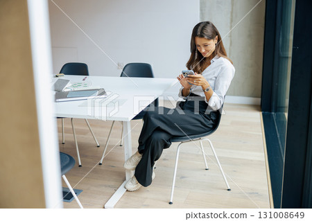 A young woman fully engaged with her smartphone while seated in a modern workspace environment 131008649